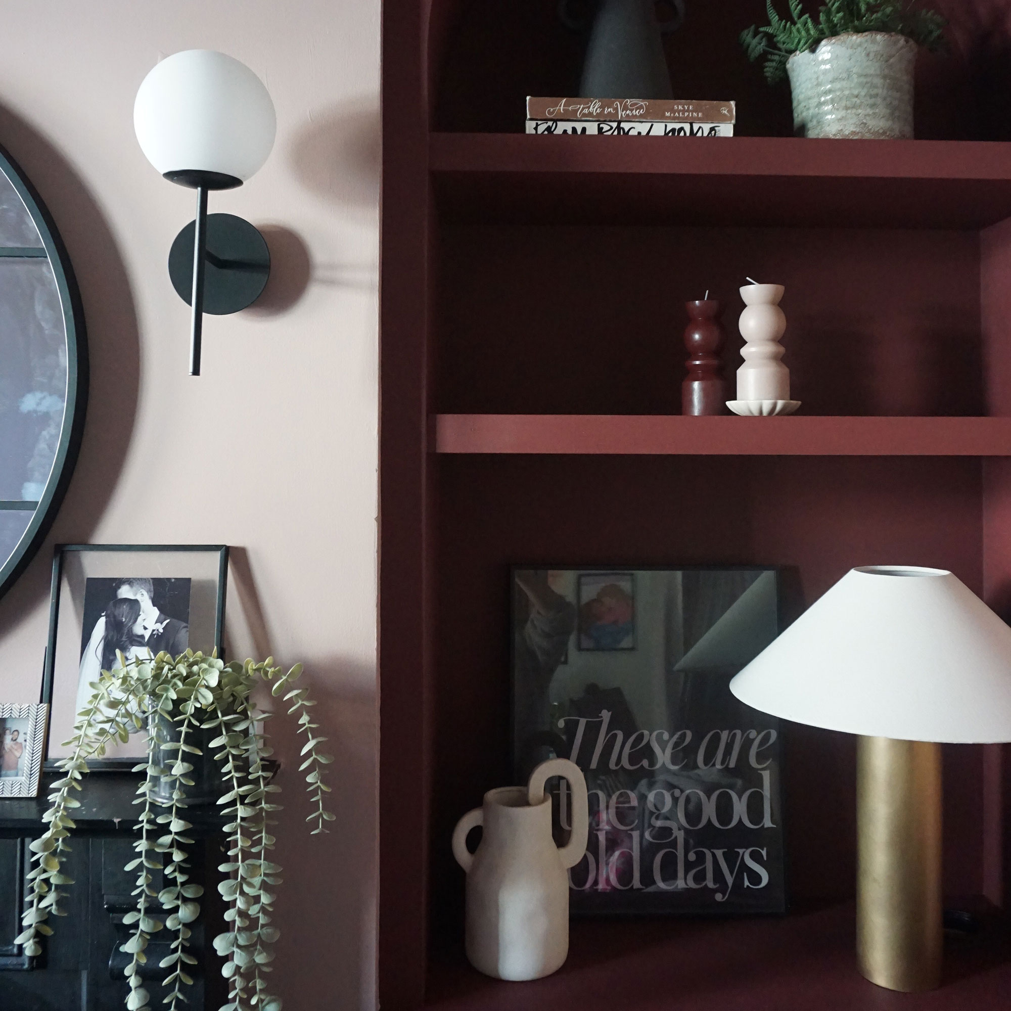 Built in shelving painted deep red, holding books, lamp and vases, beside neutral wall with wall lamp and plant on mantle