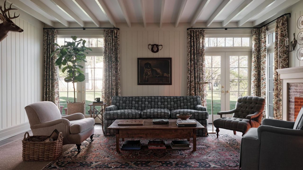 a cozy living room with rustic influence. A large, patterned sofa sits against a wall with two sets of french doors, accent chairs are on either side of the couch, a coffee table sits in the middle of the room, and a deer head hangs on the wall
