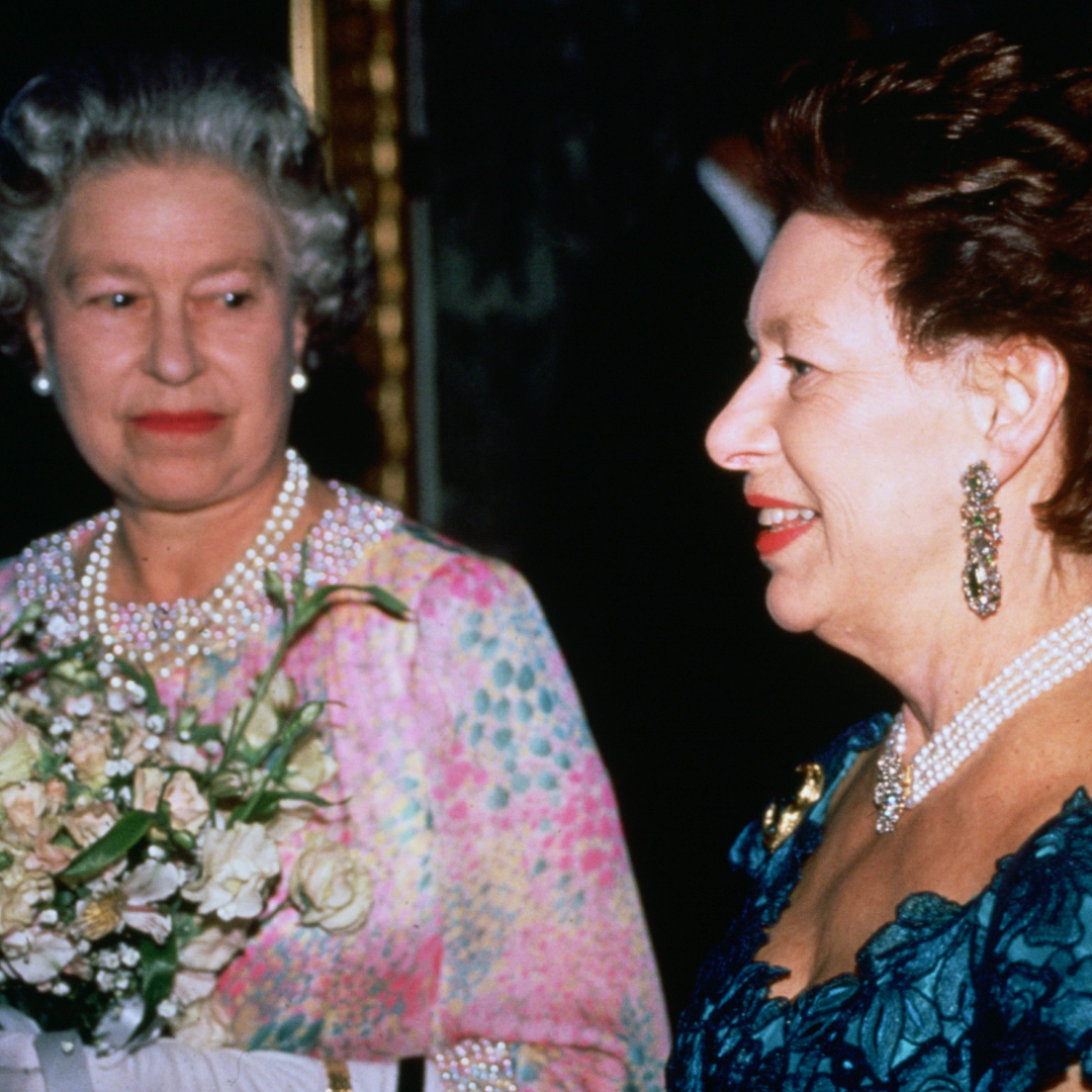Queen Elizabeth wearing a patterned dress and holding flowers smiling at Princess Margaret 