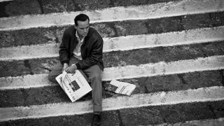 A black and white image of Andrew Scott as Tom Ripley sitting on the floor with a newspaper in his lap