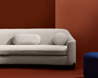View of The Drift Sofa - a grey sofa with cushions and black legs in a room with a dark terracotta floor and walls. There is also a dark blue stool beside the sofa