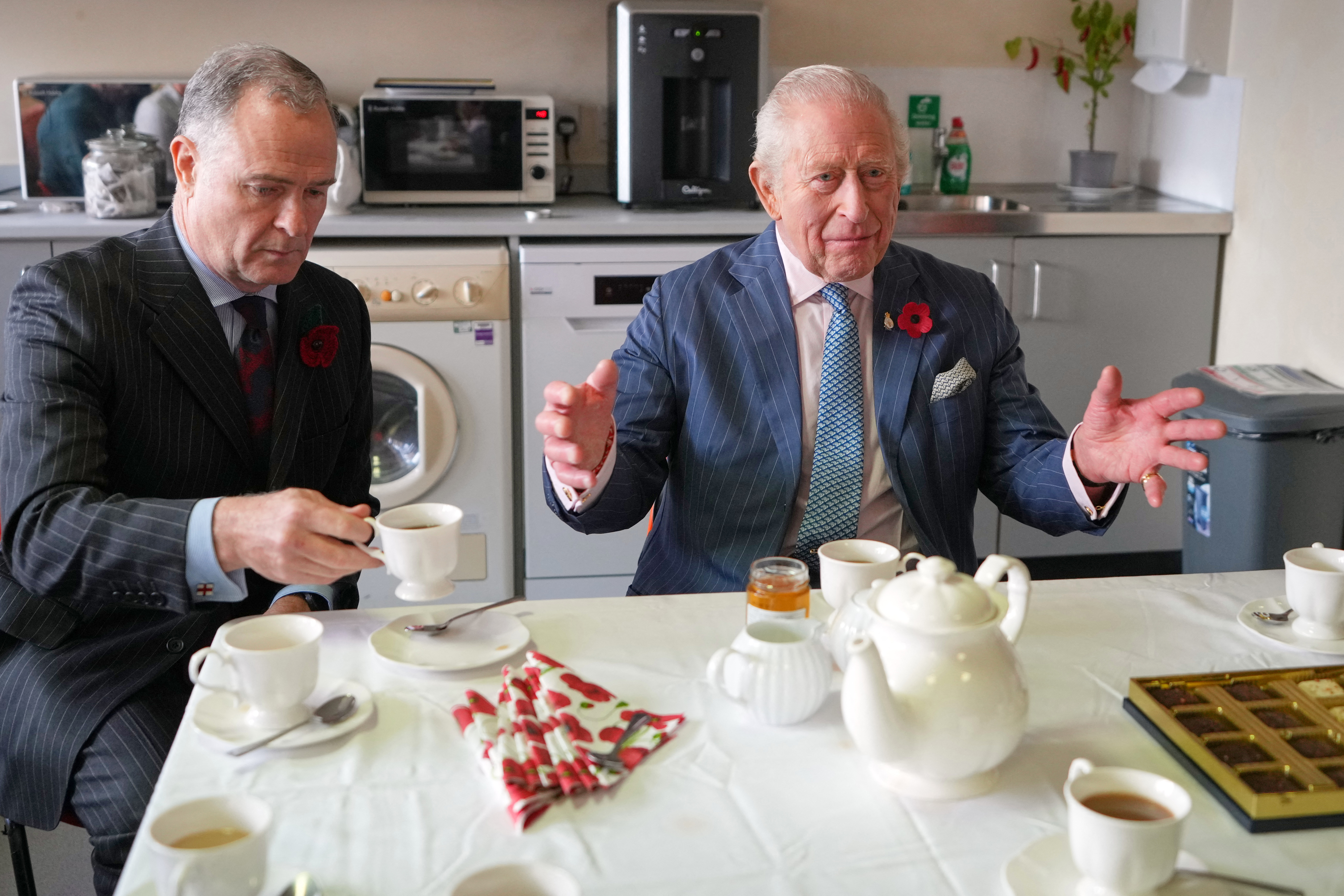 King Charles sitting at a table having tea holding his arms out
