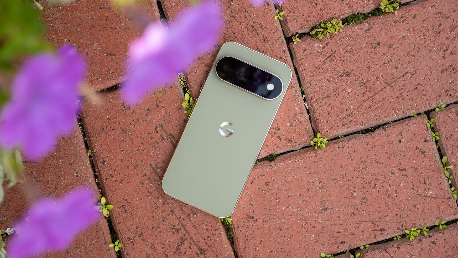 An olive green Google Pixel 11 Pro on a brick floor with purple flowers above