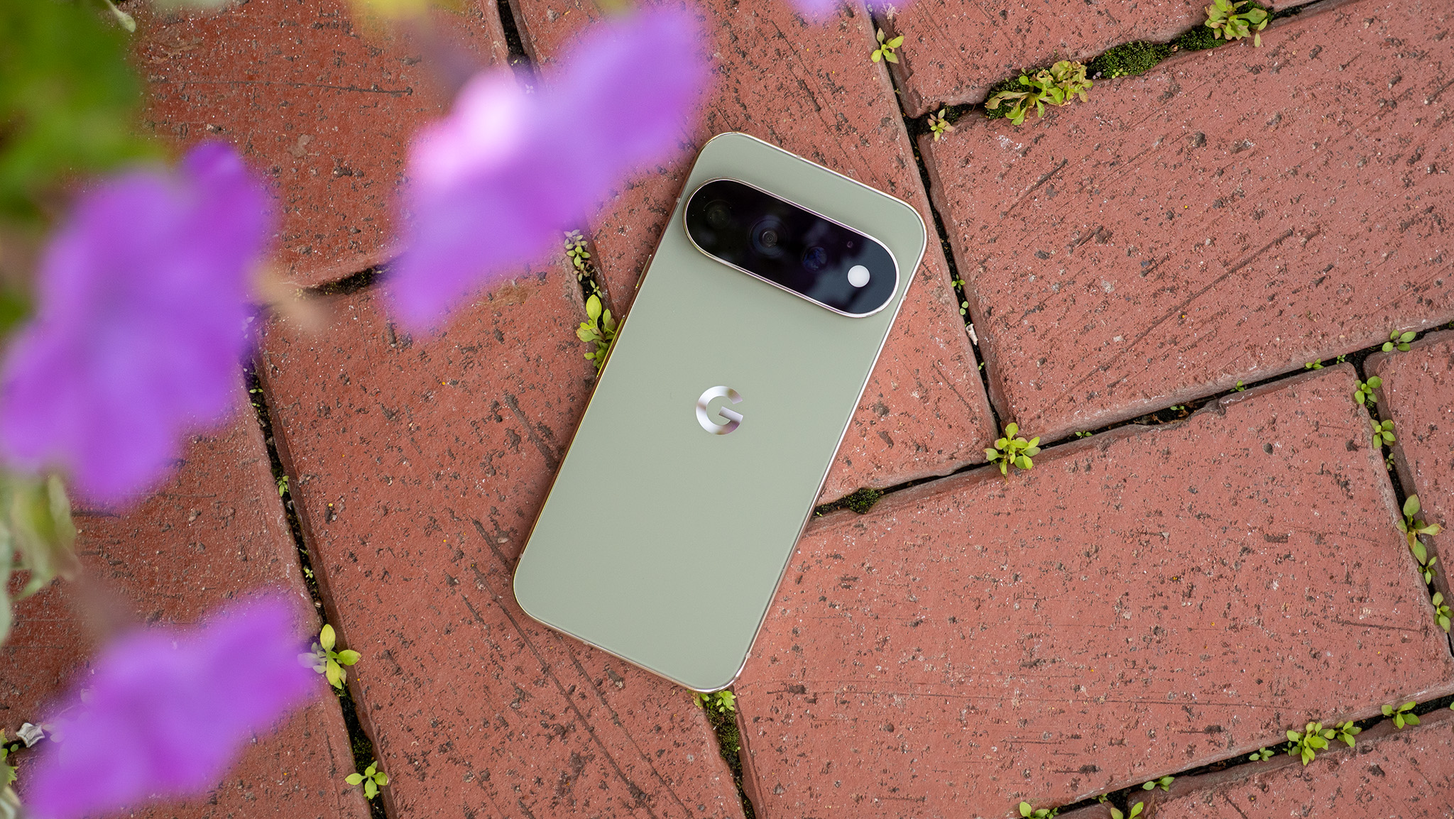 An olive green Google Pixel 11 Pro on a brick floor with purple flowers above
