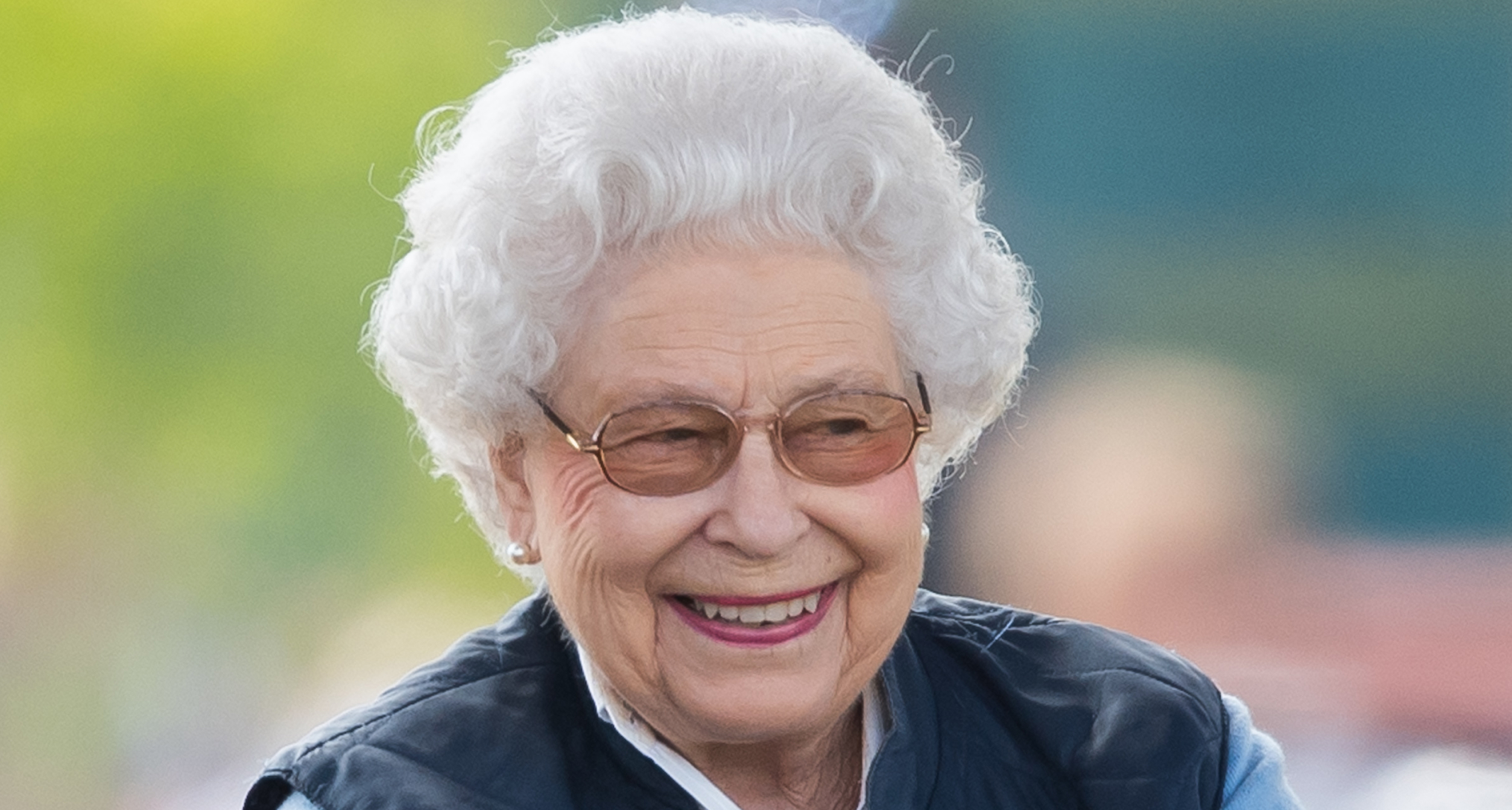 Queen Elizabeth wearing a blue vest and glasses and smiling