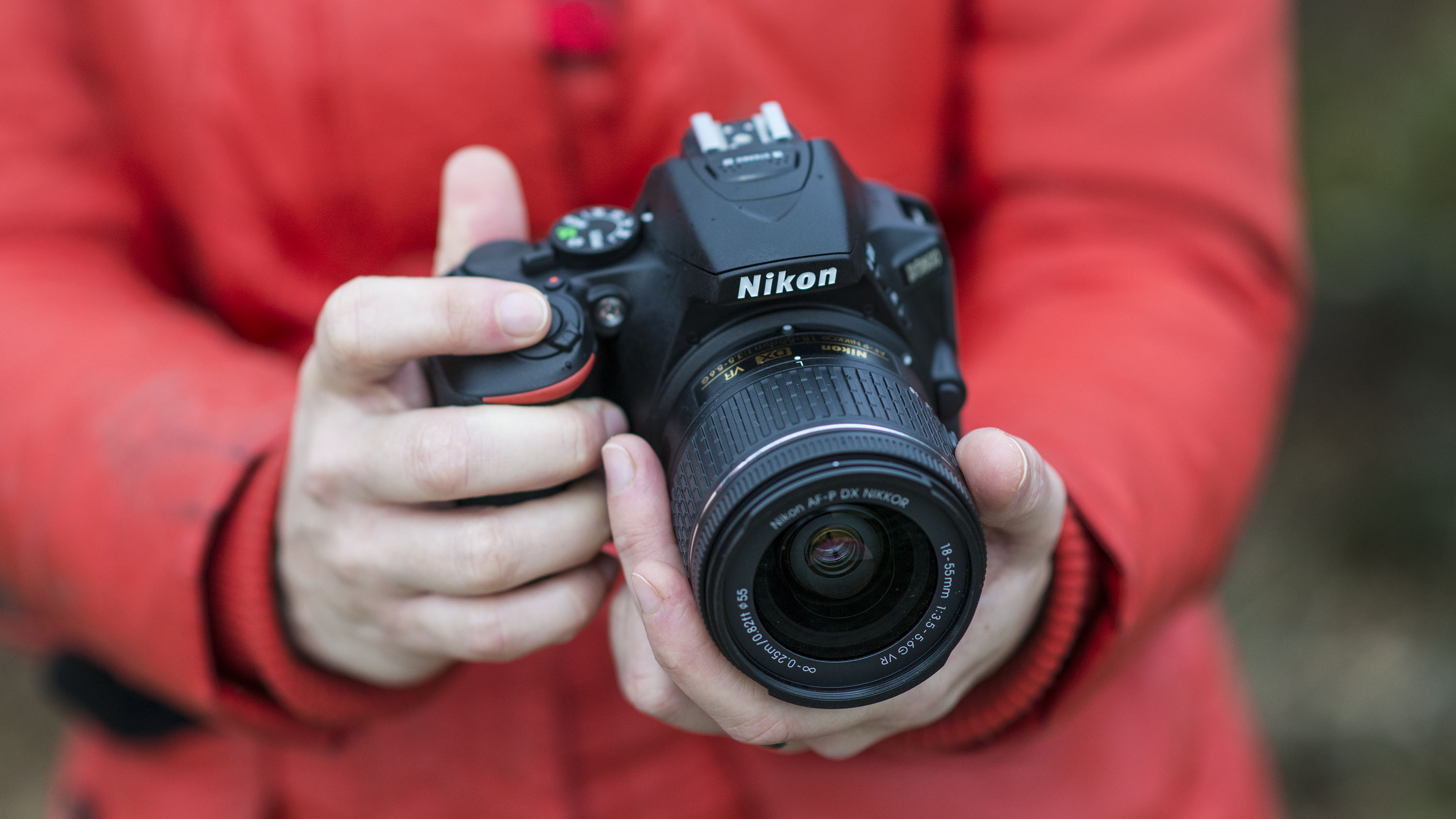 En person iklädd en röd jacka står utomhus och håller i en Nikon D5600