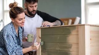 A man and woman painting a wooden chest of drawers a green colour