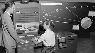 Woman sits and writes at a desk with a large panel of switches built in while a man stops to talk with her.