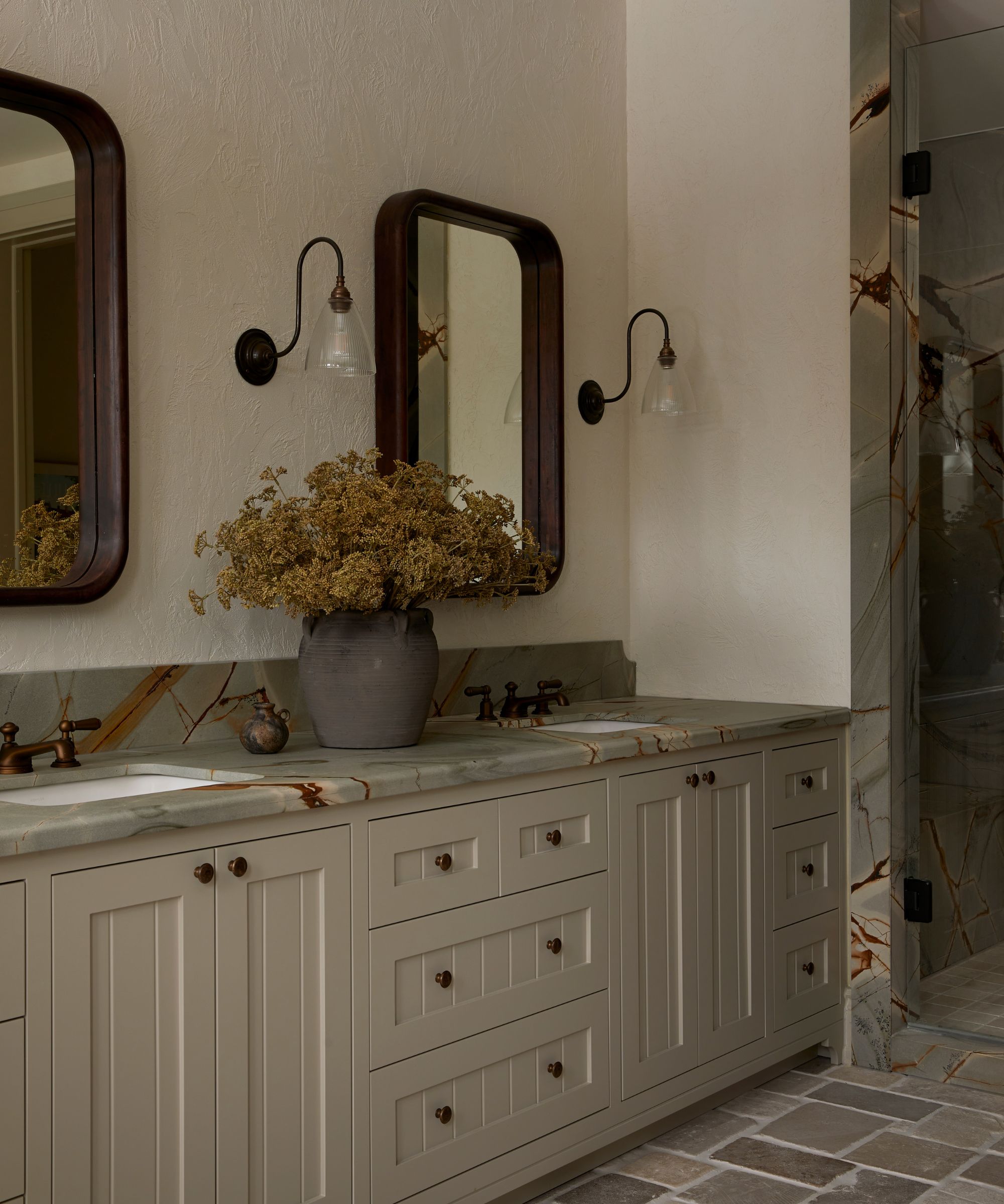 Neutral bathroom with double vanity with marble countertop, two mirrors and wall lights, vase of flowers on countertop