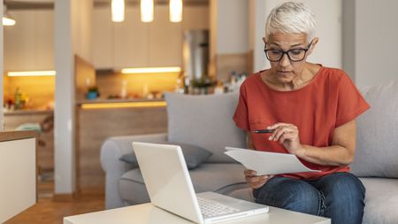 An older woman looks over some paperwork while sitting on her sofa in front of her laptop.