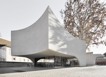 Large concrete building with curved sides, a tourist office on the ground with a tall tree behind