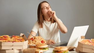 A woman sitting at a desk with a laptop open surrounded by junk food (burgers, pizzas, spaghetti)