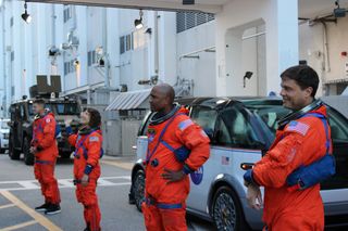 four humans standing in front of a van wearing orange spacesuits