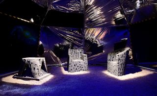 Futuristic image, moon terrain, inside view of an open tent on wheels, lights on inside, stone seating, black background, projection of a galaxy in the distance on the left