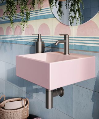 A compact bathroom with a playful aesthetic, featuring a pastel pink square wall-mounted basin with a matte black tap and matching exposed pipework. The walls are adorned with a mix of blue tiles and decorative tiles showcasing geometric patterns in soft pink, blue, and cream tones.