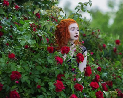 Young woman dressed in goth style stands amid a red rose bush