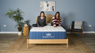 Two of our mattress testers sitting on the navy and white Bear Elite Hybrid mattress on wooden bedframe in sleep studio with blue walls, wooden bedside table, plant and black chair.