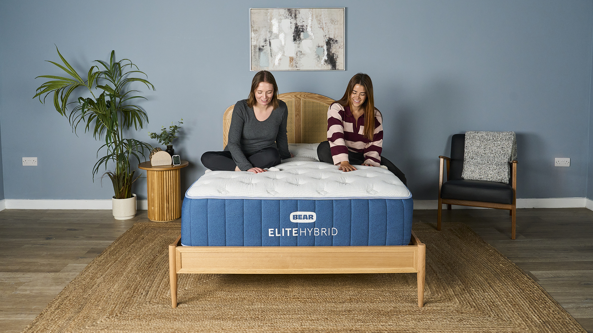 Two of our mattress testers sitting on the navy and white Bear Elite Hybrid mattress on wooden bedframe in sleep studio with blue walls, wooden bedside table, plant and black chair.