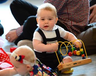 Baby Prince George holding a wooden bead toy and crawling on a floor in blue overalls