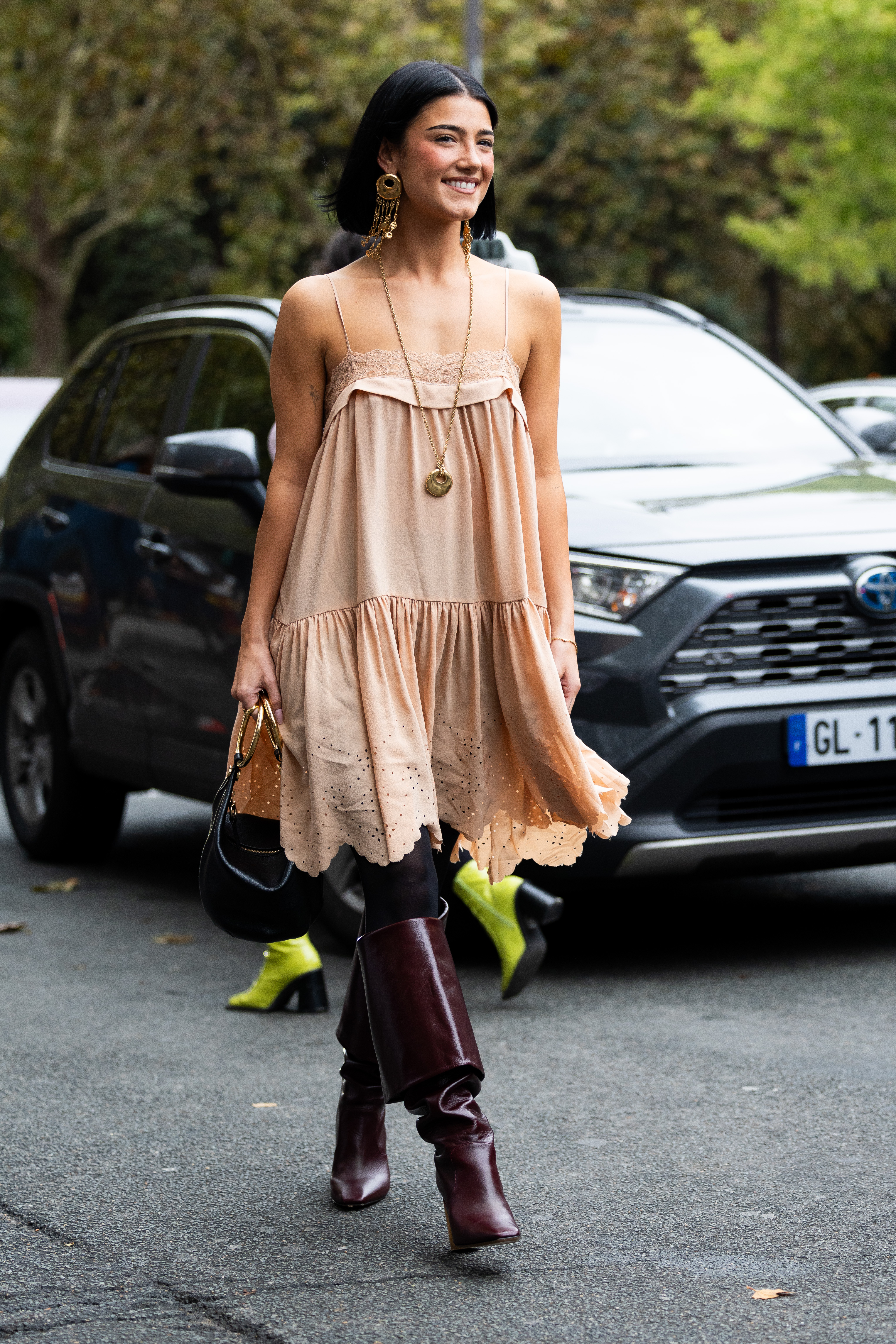 Charli D'Amelio lleva botas de tacón alto hasta la rodilla de cuero burdeos, medias negras, un bolso de cuero dorado y burdeos, pendientes colgantes dorados, un collar dorado y un minivestido corto con volantes de color rosa salmón en el exterior del desfile de Chloé durante la Semana de la Moda de Mujer Primavera/Verano 2025 como parte de la Semana de la Moda de París el 9 de septiembre.