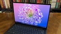 A laptop on a wooden table displays a vibrant, colorful chrysanthemum on its screen. Background: a bookshelf lined with assorted books. Cozy ambiance.