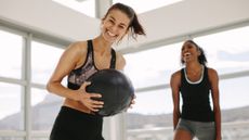Woman in fitness studio holding medicine ball and smiling, another woman in the background is laughing