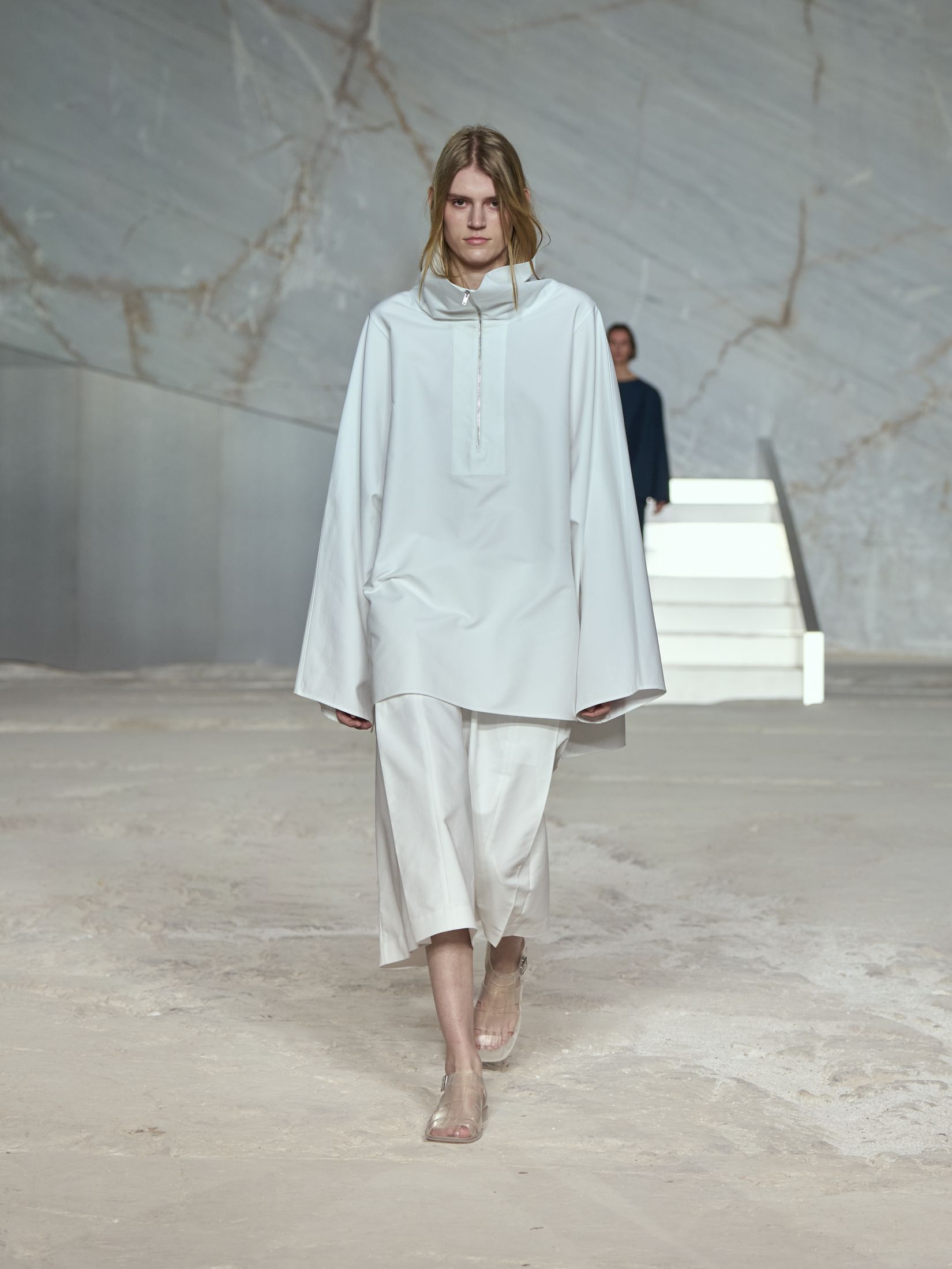 COS model wearing a white sporty poplin anorak with long culotte shorts and clear sandals at the S/S 25 show in Greece.