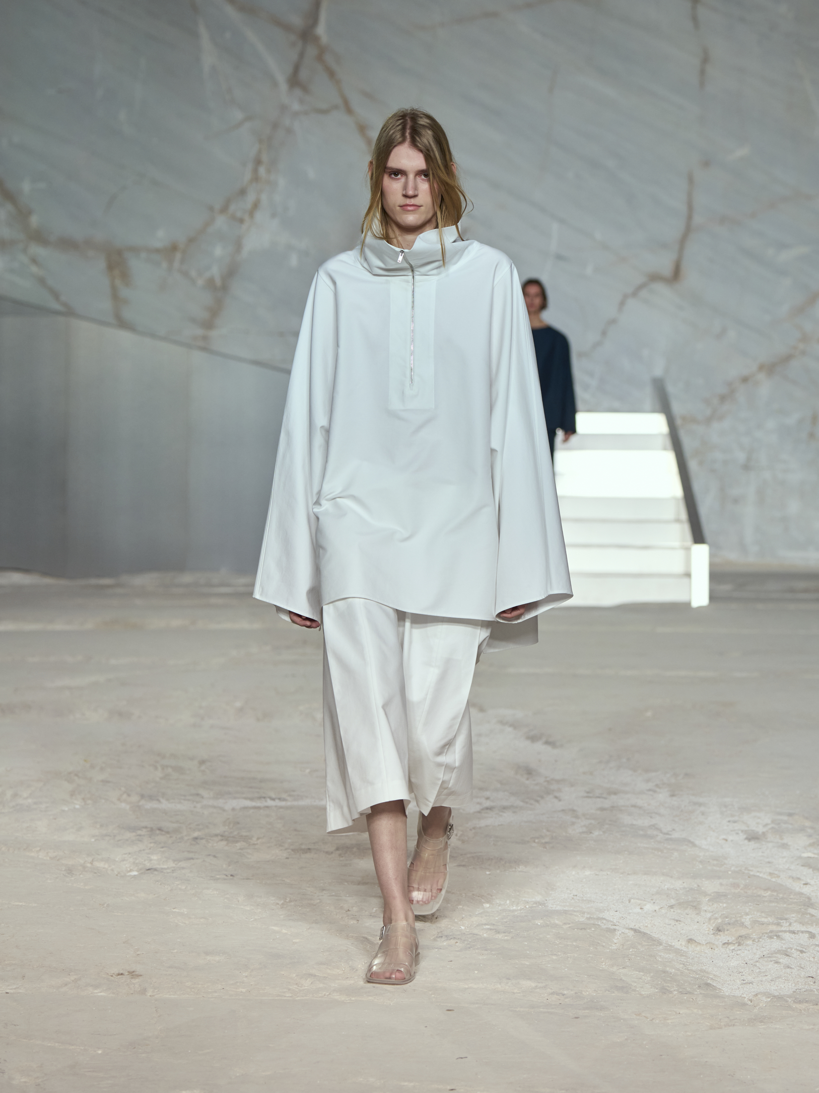 COS model wearing a white sporty poplin anorak with long culotte shorts and clear sandals at the S/S 25 show in Greece.