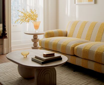A yellow themed living room with a striped sofa, a coffee table, and a vase of yellow mimosa flowers on a pedestal 