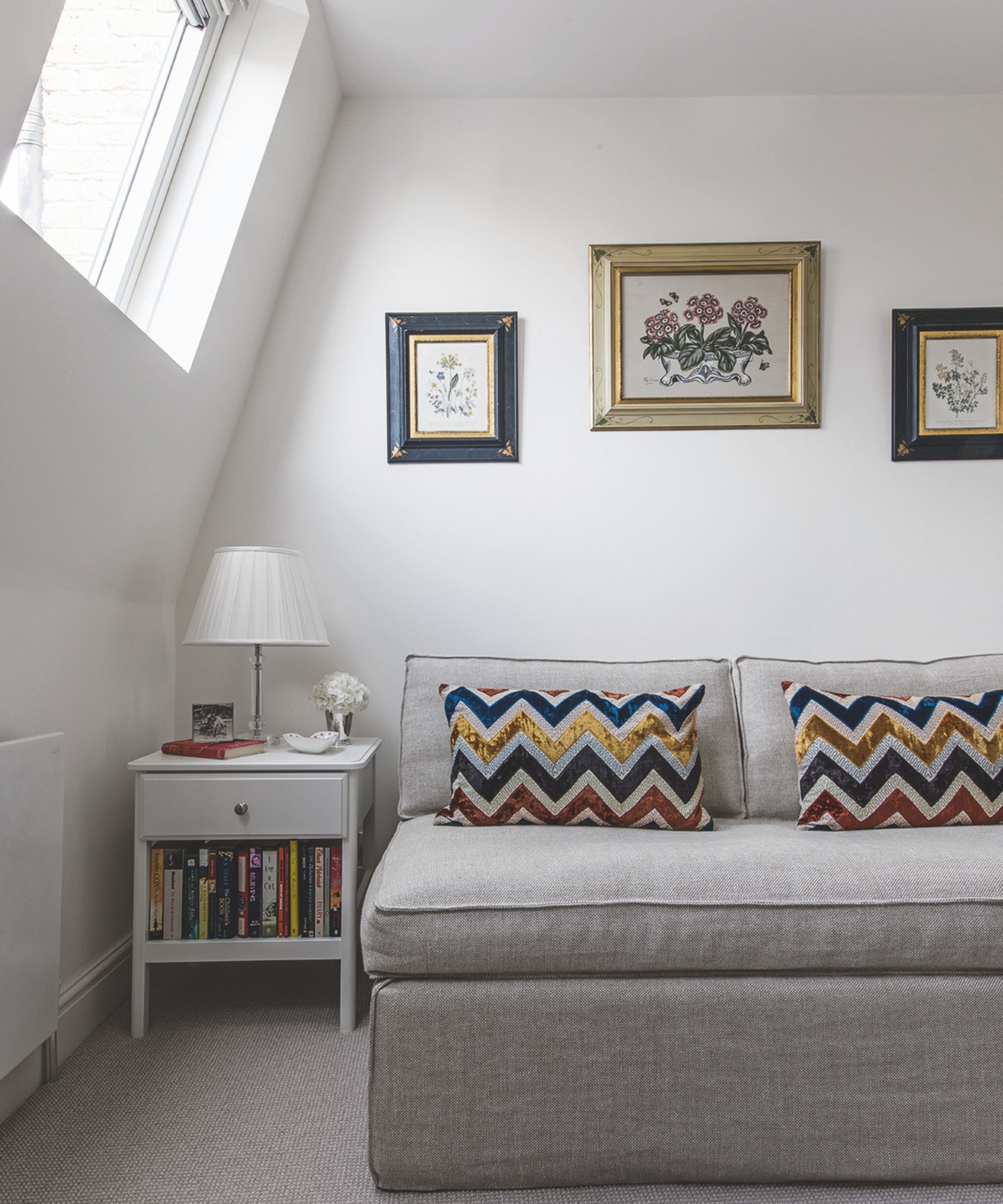 An attic seating area with a grey couch, chevron patterned cushions, white bedside table filled with books, white lamp and framed artwork above.