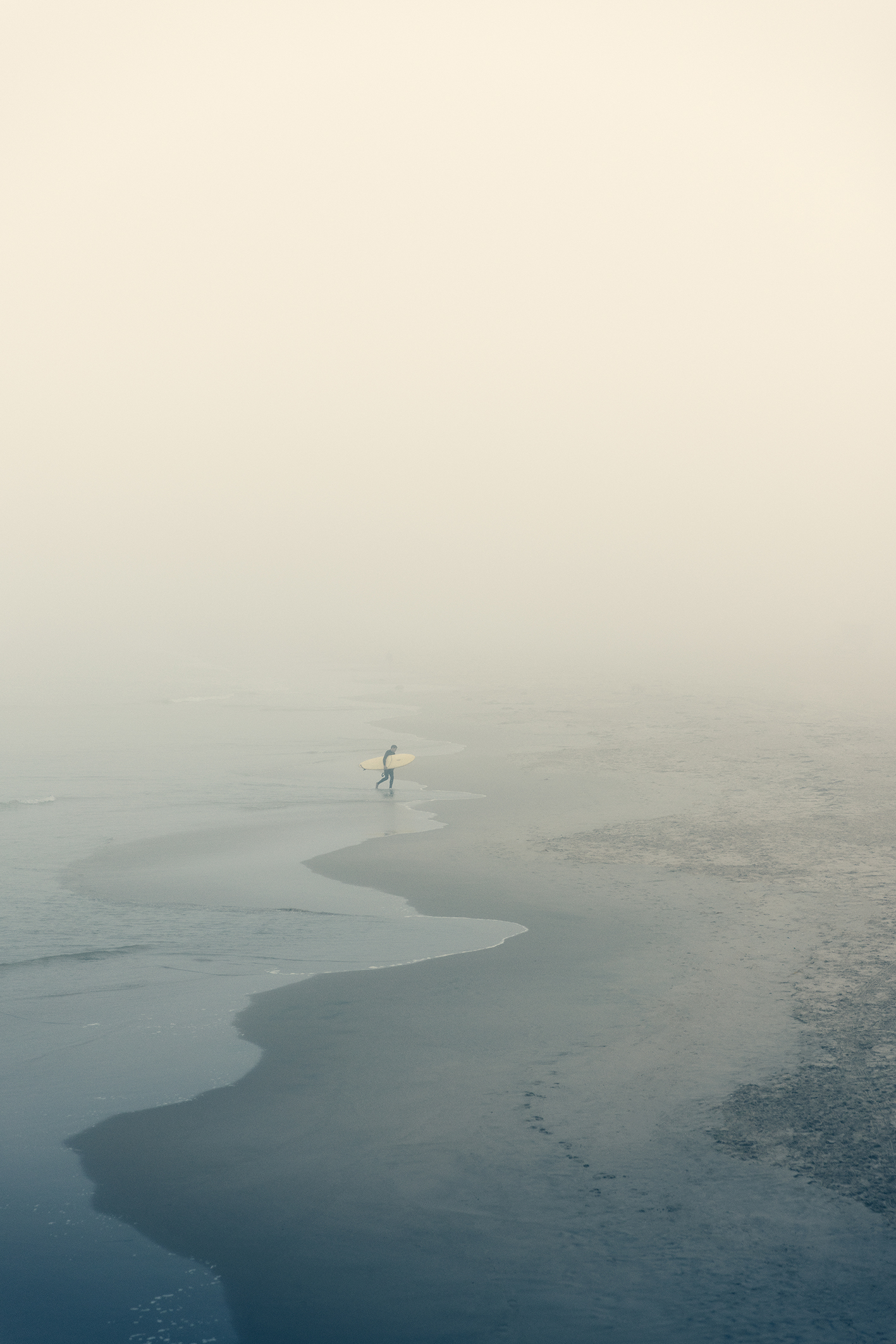 A solitary surfer with a yellow board walks along a misty beach, with soft waves lapping at the shore