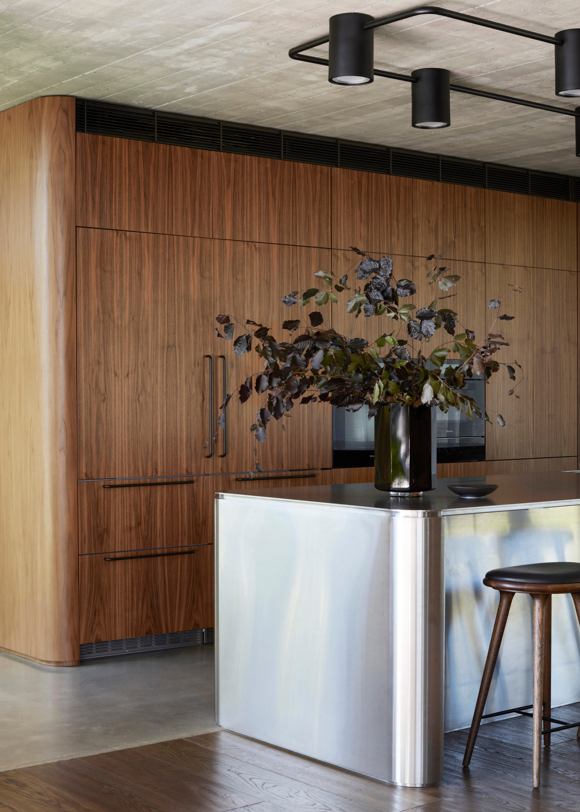 A kitchen with wood and stone flooring, wood cabinetry, a metallic island with dark wood stools and a vase of stems