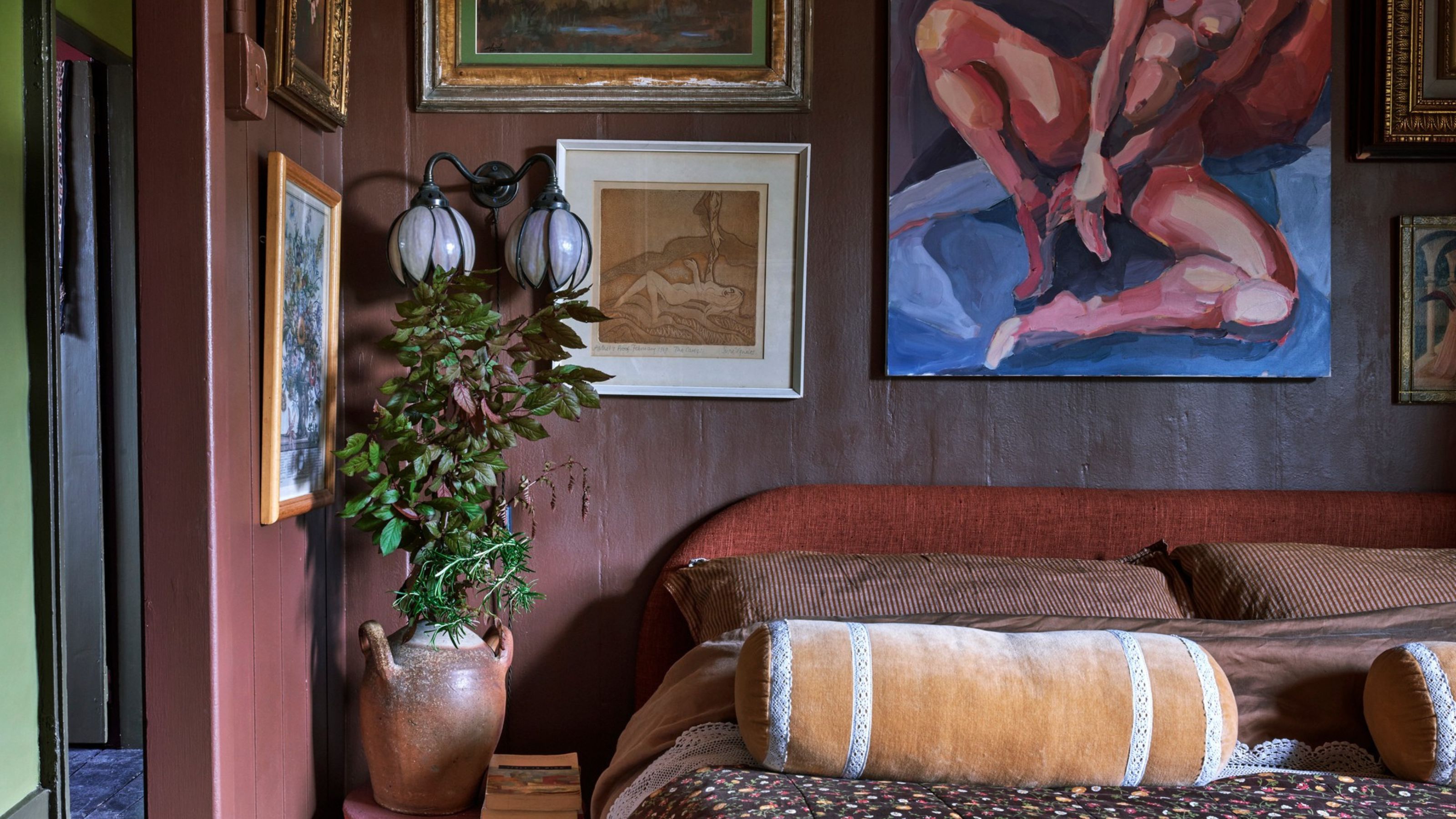bedroom with earthy red walls and gallery wall of art with terracotta urn with branches and yellow bolster cushion