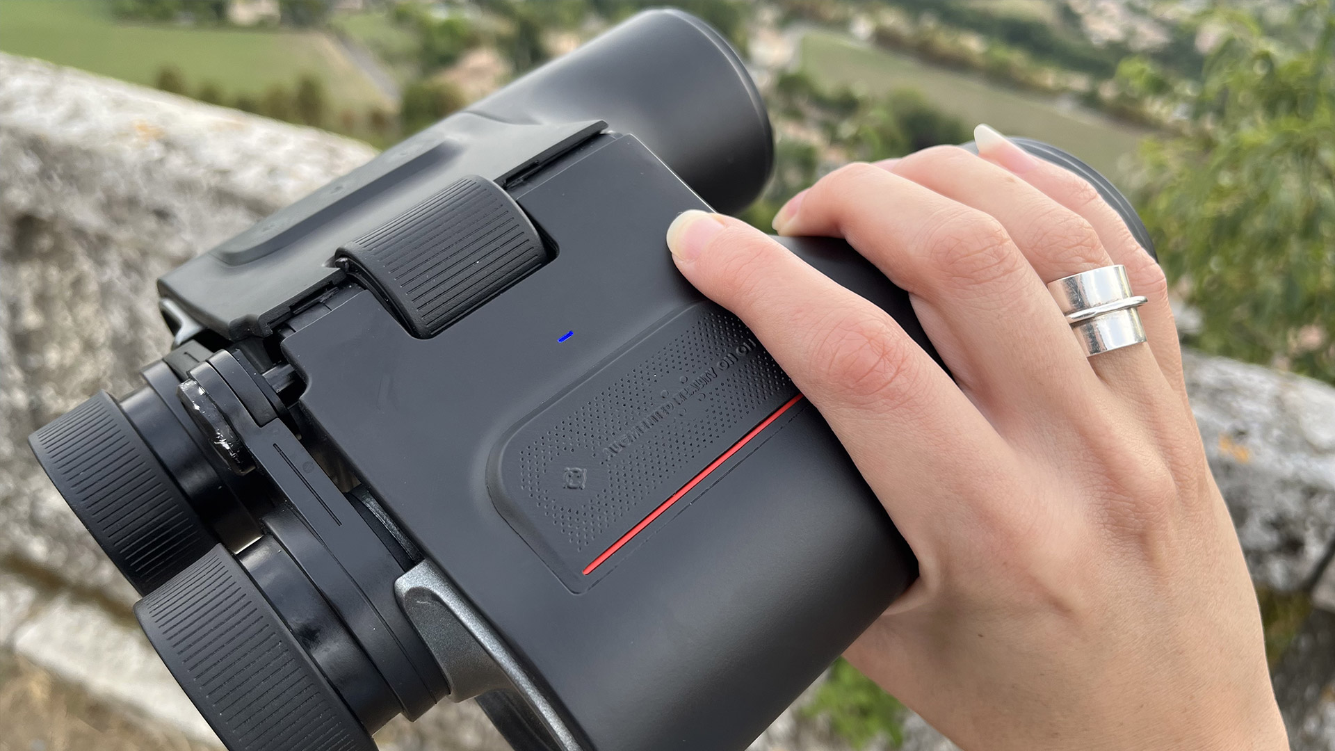 A hand holds a pair of black binoculars above a stone handrail