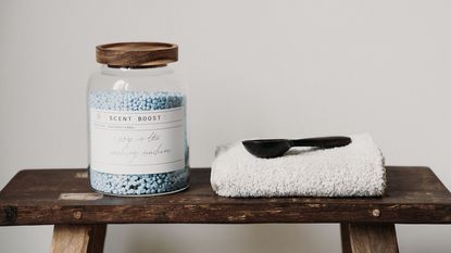 A glass jar with a wooden lid full of laundry beads, a folded towel and a scoop beside it, on a wooden table
