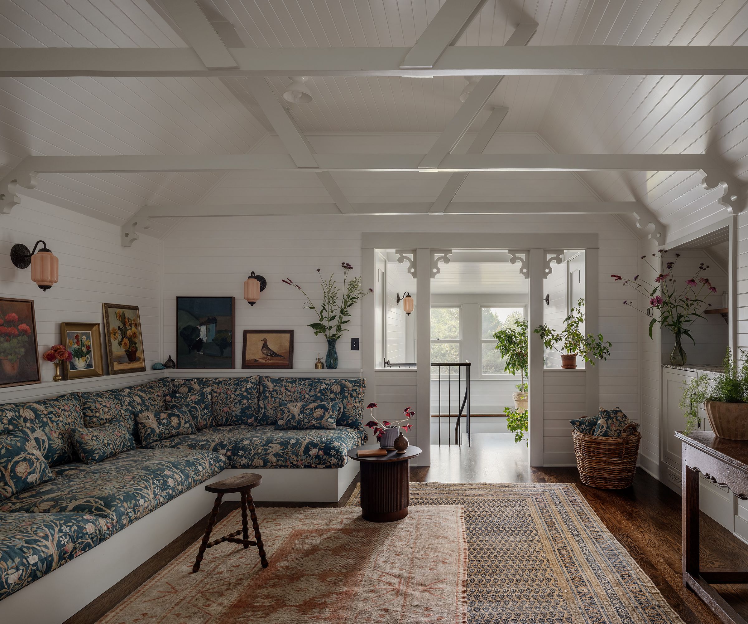 Living room with white walls and beamed ceiling, built-in banquette with floral cushions, picture ledge with vintage paintings and large Persian rug
