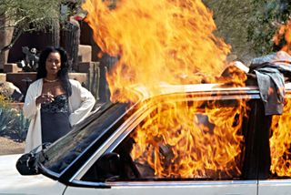 A woman smokes a cigarette as she watches a car burn.