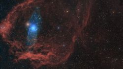 A blue glowing star is surrounded by red gas in deep space.