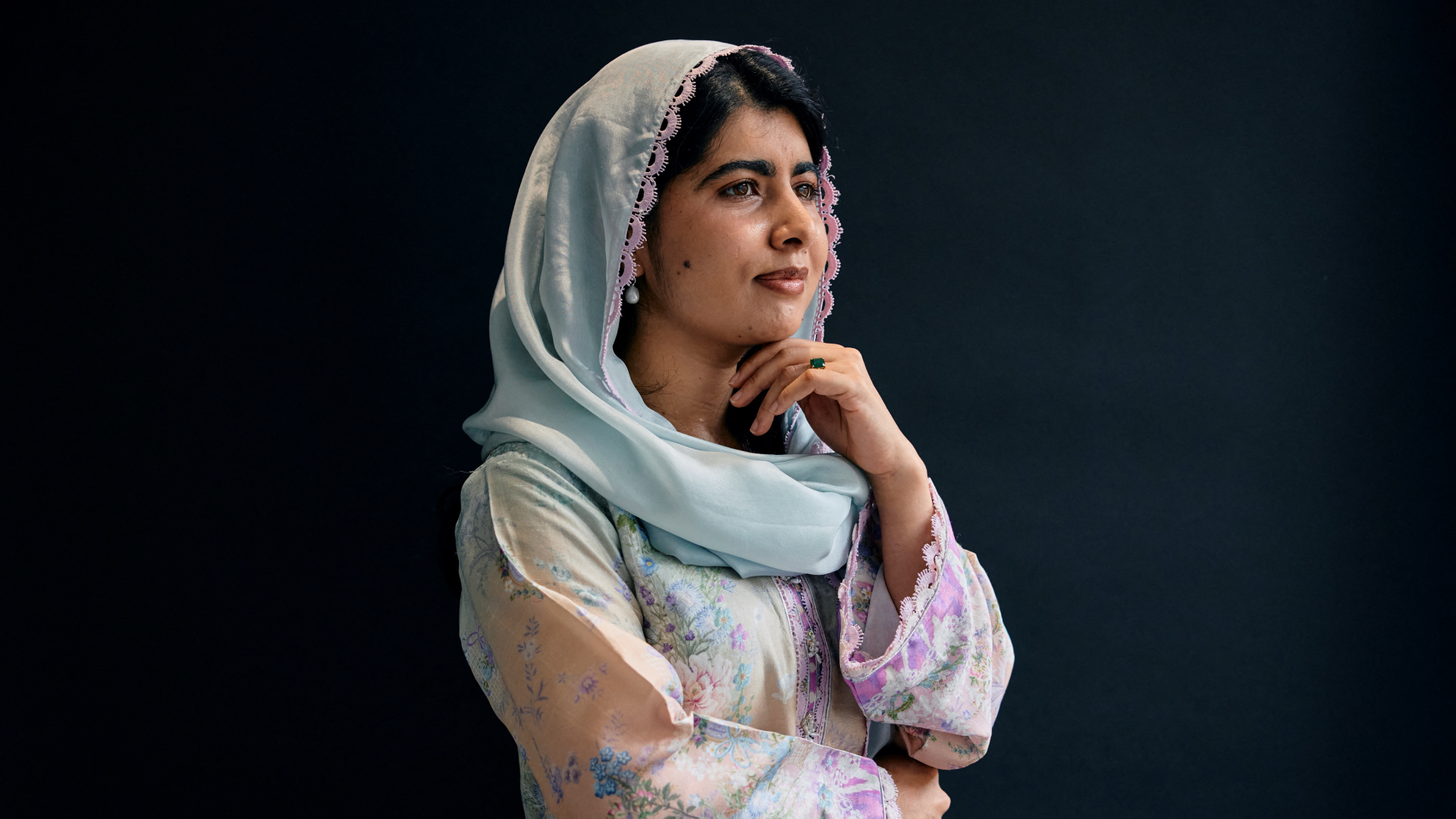 Malala Yousafzai poses in a blue headscarf and floral outfit against a dark backdrop