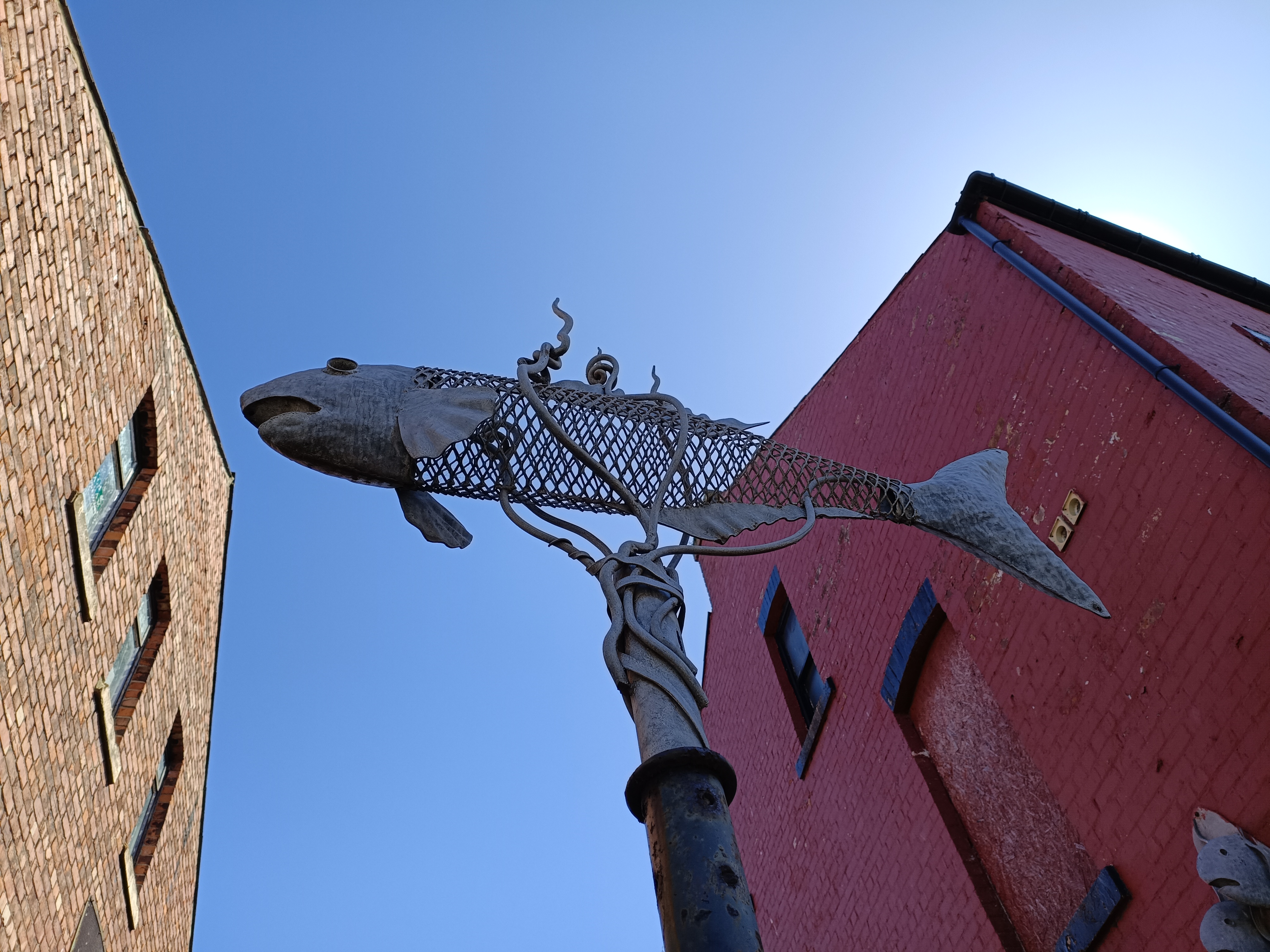 A metal sculture of a fish suspended on a pole beneath a blue sky