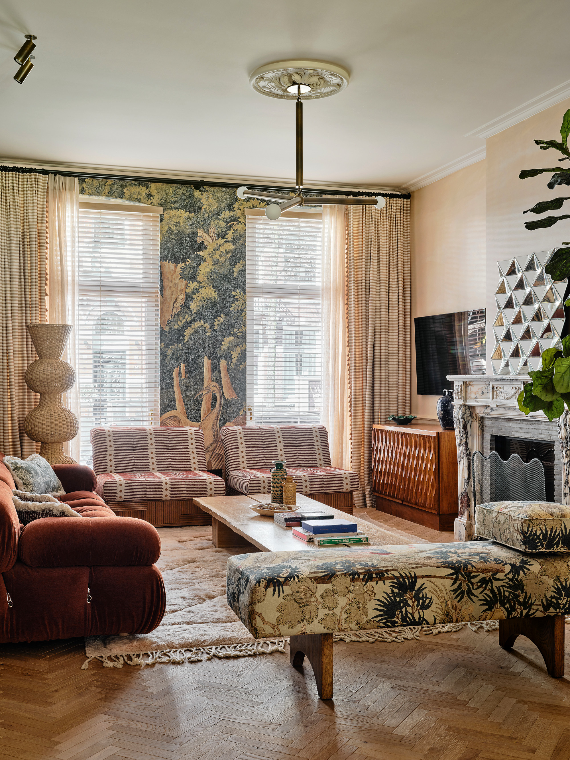 Living room with botanical mural and apricot walls, botanical upholstered bench, rust-colored Camaleonda sofa, striped side chairs, wood coffee table and peach geometric rug