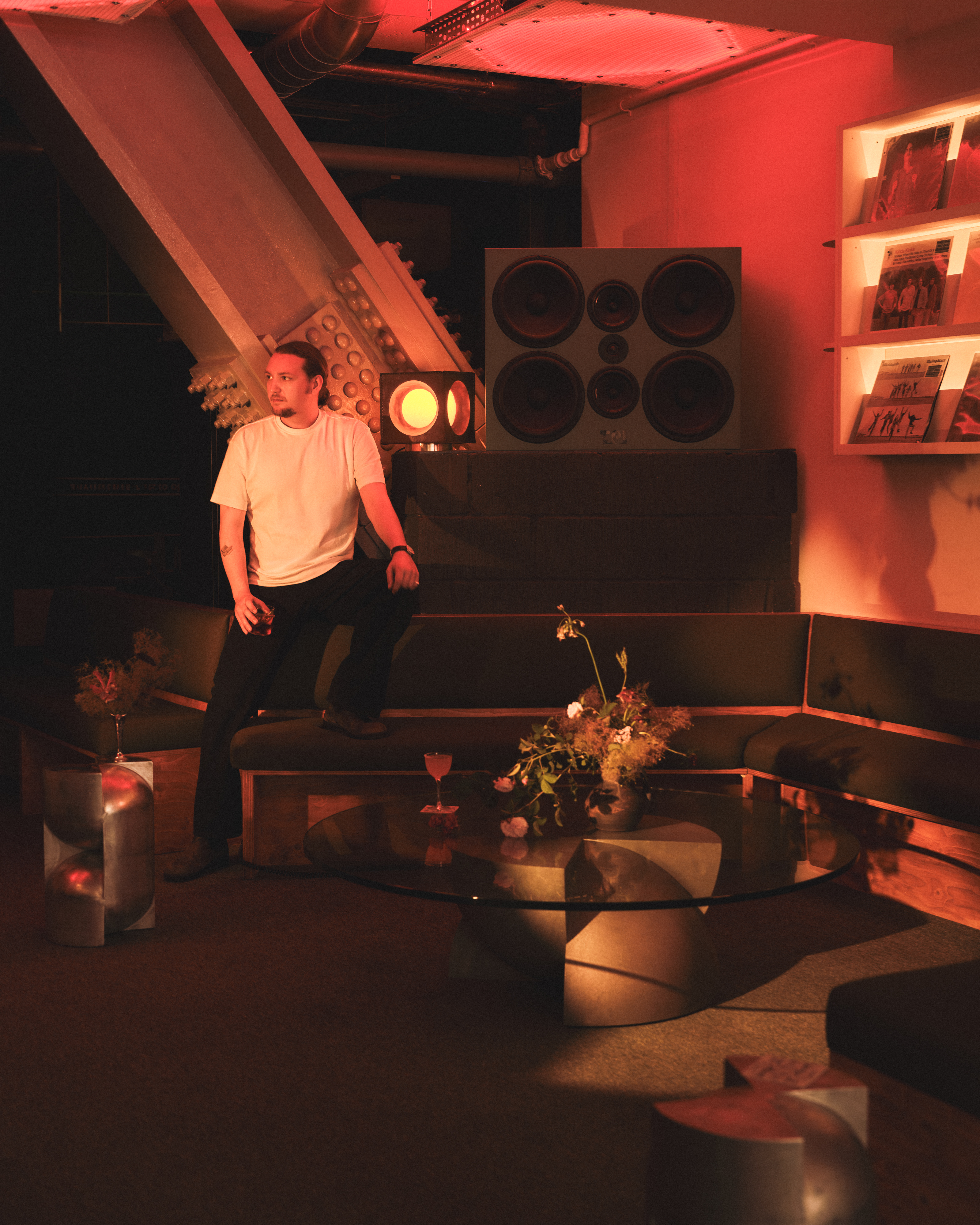 A man dressed in a white T-shirt and black trousers poses sideways while leaning on a wood and fabric banquette surrounded by hi-fi music gear and retro-futuristic furniture.