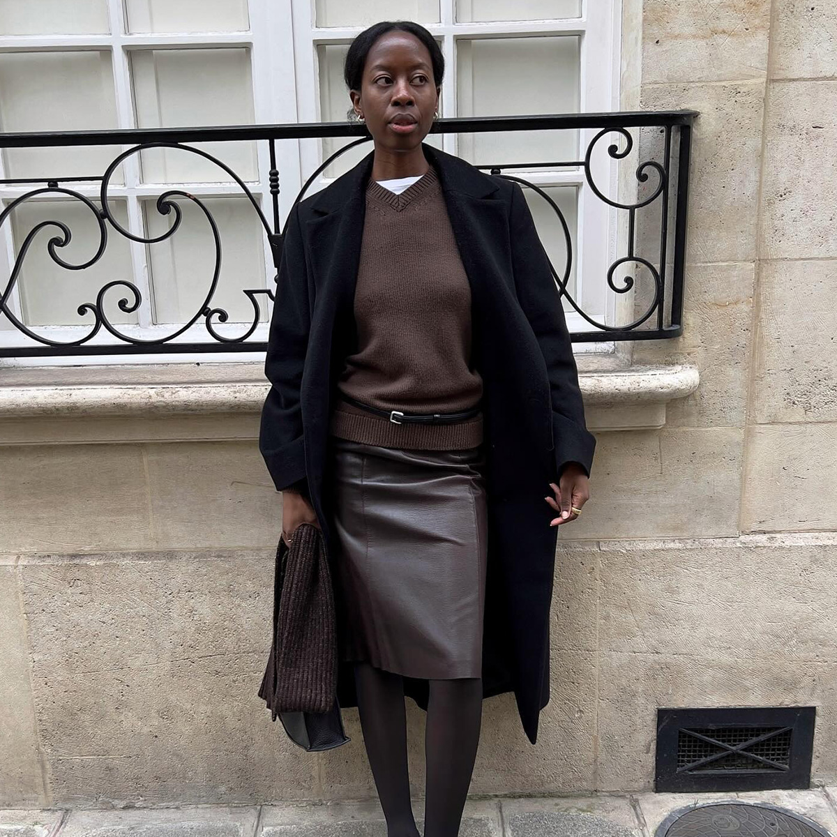 Sylvie wears a black coat, brown v-neck sweater, and brown skirt.