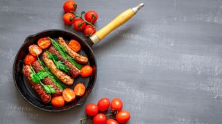 A cast iron skillet with a wooden handle containing 5 sausages and halved tomatoes