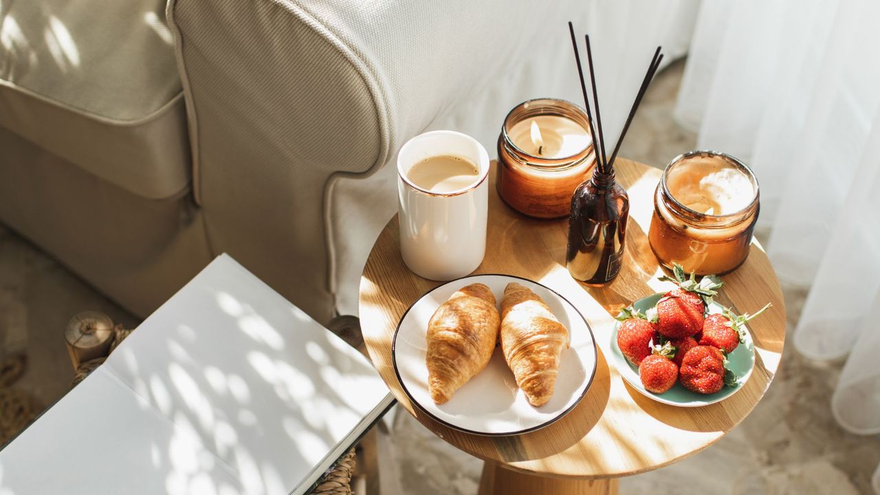 A reed diffuser on a side table with candles and croissants next to an arm chair