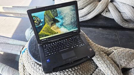 A rugged laptop with a seashore wallpaper is placed on thick, coiled ropes, suggesting durability and adventure. Sunlight casts soft shadows on the scene.