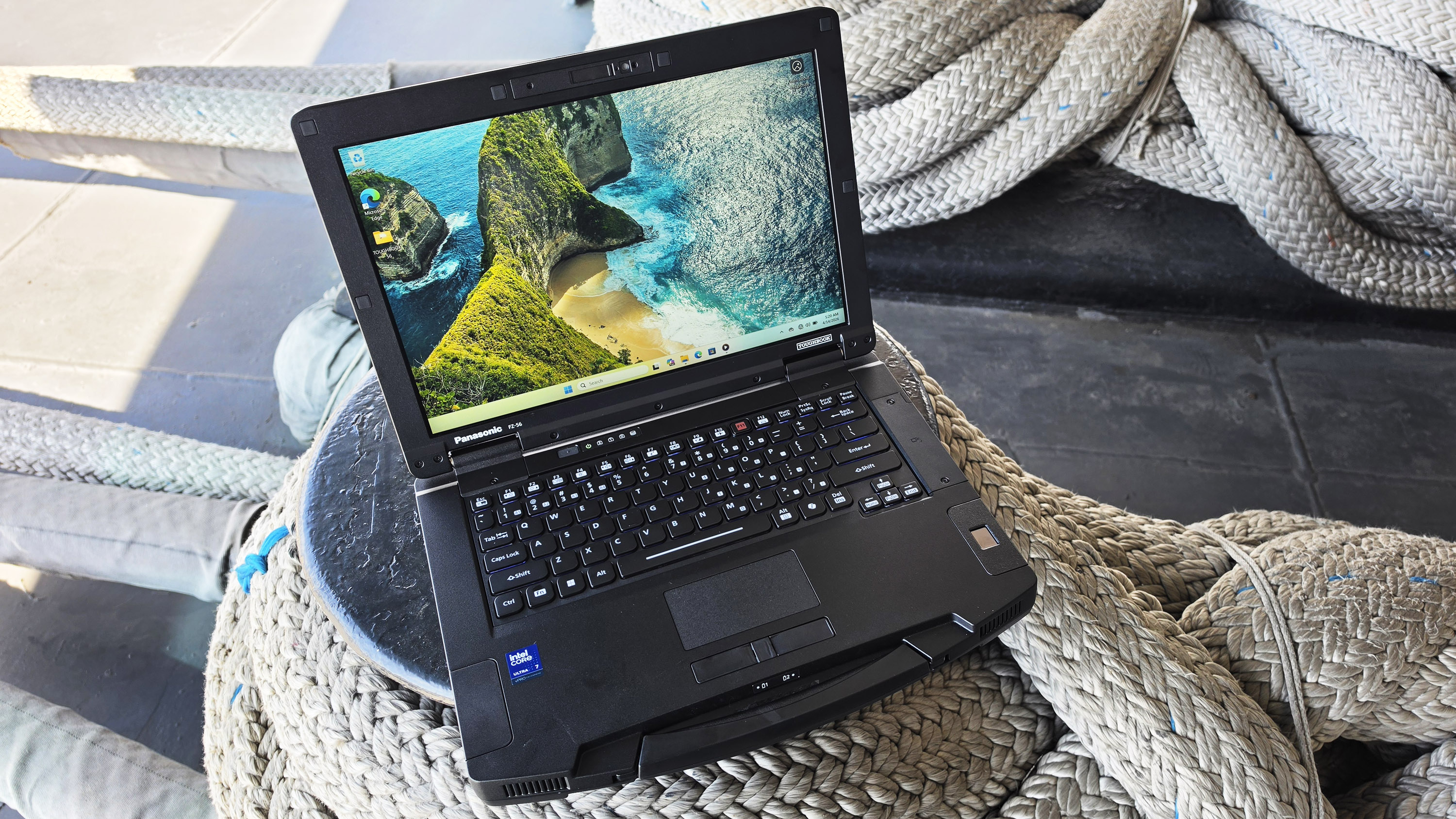 A rugged laptop with a seashore wallpaper is placed on thick, coiled ropes, suggesting durability and adventure. Sunlight casts soft shadows on the scene.
