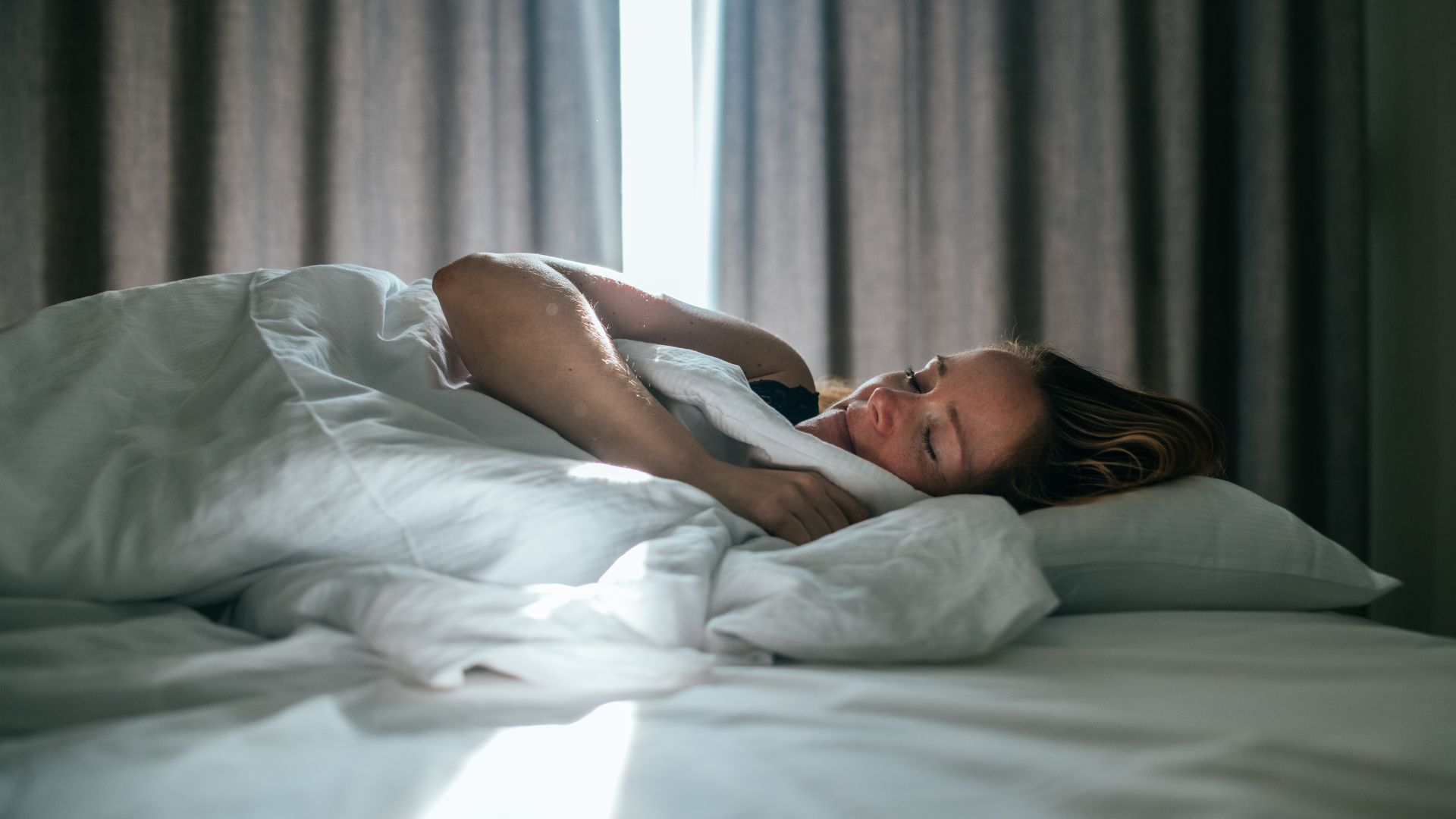 A woman lies asleep in a bed that has all white bed linen. In the background are curtains that are slightly open, letting sunlight in.
