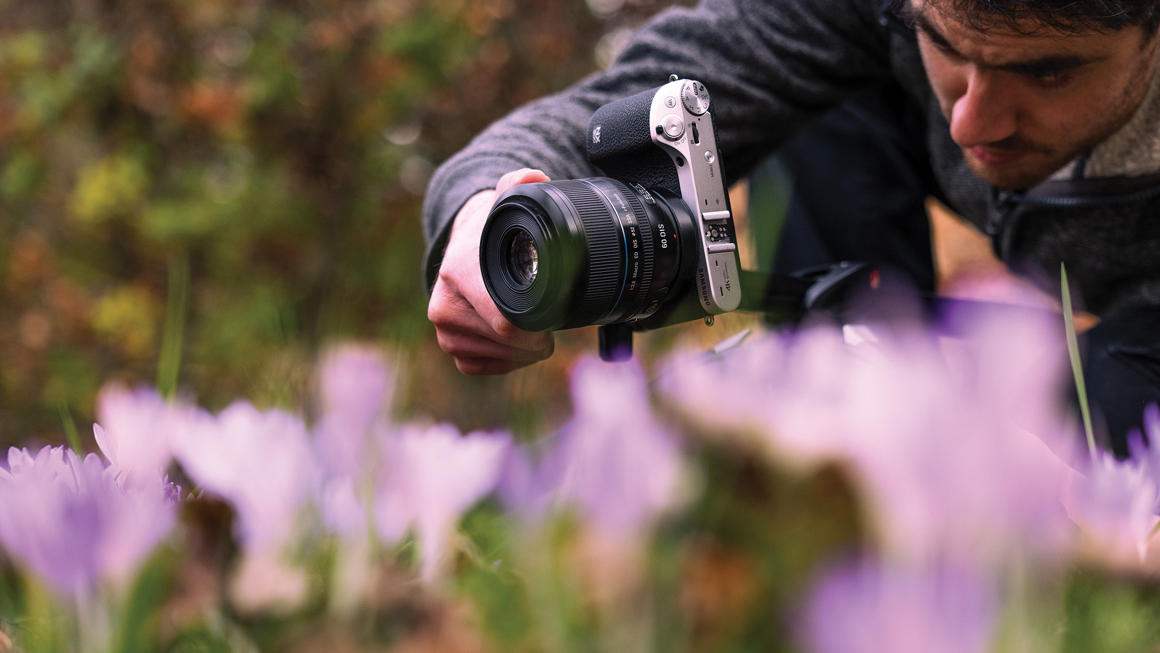 A photographer crouches, focusing a camera on purple flowers in a garden. The setting is vibrant and natural