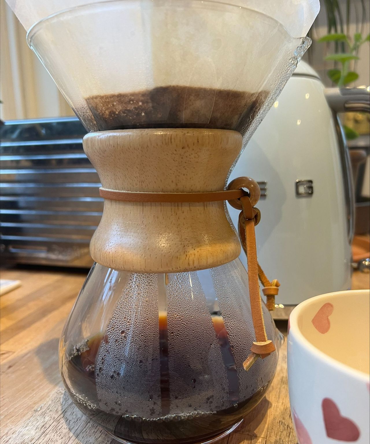 Brewing coffee on a wooden countertop with the Chemex coffeemaker. Behind is a silver Dualit toaster, a baby blue Smeg kettle, and a houseplant.